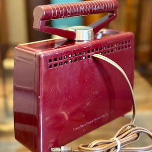 May include: A vintage burgundy-colored appliance with a handle and a dial. The appliance has a rectangular shape with ventilation holes and a power cord. The handle is a darker shade of burgundy. The text "Miracle Maid" is visible on the handle.