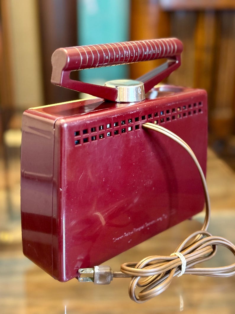 May include: A vintage burgundy-colored appliance with a handle and a dial. The appliance has a rectangular shape with ventilation holes and a power cord. The handle is a darker shade of burgundy. The text "Miracle Maid" is visible on the handle.