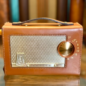 Puede incluir: Una radio Philco rectangular revestida en cuero color canela con un asa superior. La parte frontal presenta una rejilla de altavoz de malla plateada, un dial de sintonización circular dorado con números y un emblema plateado de la marca Philco.