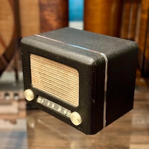 May include: A retro-style black radio with a textured finish. The radio has a tan speaker grille, a tuning dial, and two clear knobs. Silver trim accents the edges. The radio is a vintage design.