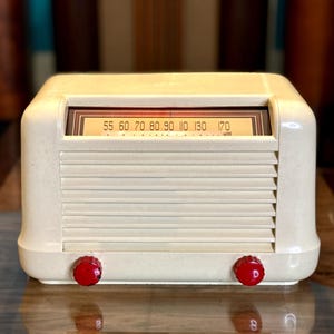 Könnte beinhalten: Ein cremefarbenes Tischradio mit einem horizontalen Lamellen-Lautsprechergitter und zwei leuchtend roten, runden Drehknöpfen an der Basis. Das Radio verfügt über eine rechteckige Skala mit schwarzen Zahlen von 55 bis 170.