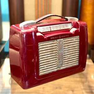 May include: A deep red General Electric portable radio featuring a glossy finish, a top carrying handle, and a silver-toned horizontal speaker grille. The radio includes a white frequency dial with numbers ranging from 55 to 160 and side control knobs.