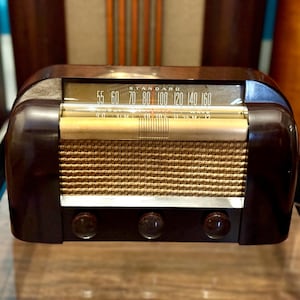 Könnte beinhalten: Ein Vintage-Tischradio mit einem dunkelbraunen, abgerundeten Kunststoffgehäuse und einem goldfarbenen Stofflautsprechergitter. Die Vorderseite zeigt eine horizontale Glasskala mit dem Wort STANDARD über Frequenzzahlen und drei runde Knöpfe.