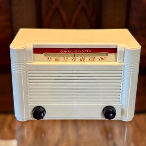 May include: A cream-coloured General Electric radio with a rectangular body, horizontal speaker grille, and two dark brown circular knobs. The radio features a red horizontal display panel with the text General Electric and frequency numbers 55 to 160.