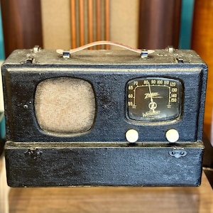 May include: A black portable Zenith Trans-Oceanic radio with a textured exterior, a top carrying handle, a speaker grille on the left, and a glass-covered tuning dial on the right. Two white circular knobs are positioned below the dial.
