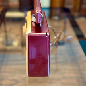 May include: A vintage radio with a burgundy and cream-colored plastic casing. The radio has a burgundy handle and a dial on the side. A power cord extends from the back. The radio is likely from the mid-20th century.
