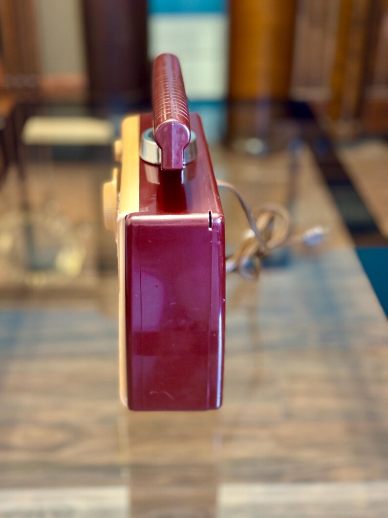 May include: A vintage radio with a burgundy and cream-colored plastic casing. The radio has a burgundy handle and a dial on the side. A power cord extends from the back. The radio is likely from the mid-20th century.