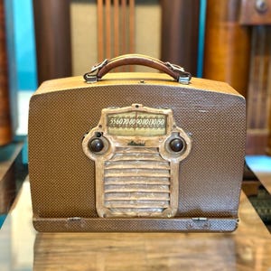 May include: A portable brown radio with a textured exterior, a leather-like handle, and a central control panel featuring a horizontal frequency dial marked from 55 to 150, two circular knobs, and a slatted speaker grille with a small logo.