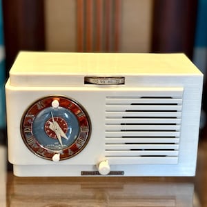 May include: A white General Electric radio with a rectangular body and horizontal speaker slats. It features a circular analogue clock face with a blue centre, white hands, and Roman numerals, alongside a horizontal radio frequency tuning dial.