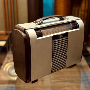 May include: A vintage radio with a cream-coloured body, dark brown wooden accents, and a brown handle. The radio features a speaker grill and a clear dial. A classic piece of electronics.