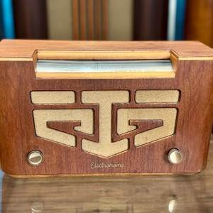 May include: A rectangular wooden radio with a warm brown finish, featuring a central speaker grille with a textured beige fabric inlay. The front panel displays the Electrohome brand name in script, two circular metallic knobs, and a top dial.