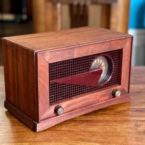 May include: A rectangular wooden radio with a dark mahogany finish, featuring a dark red grid speaker cover, a circular analogue frequency dial, and a prominent triangular pointer. Two small circular control knobs are positioned at the bottom.