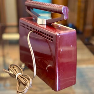 May include: A vintage burgundy-colored electric travel iron with a retractable handle. The iron has a silver-colored dial and a series of ventilation slots. A brown power cord is coiled next to the iron.