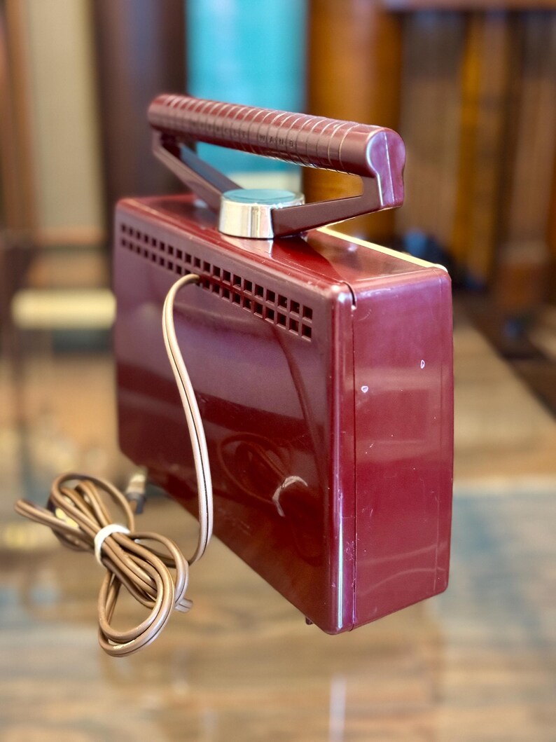 May include: A vintage burgundy-colored electric travel iron with a retractable handle. The iron has a silver-colored dial and a series of ventilation slots. A brown power cord is coiled next to the iron.