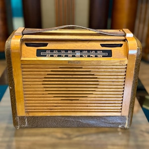 May include: A portable Philco radio featuring a light wood-grain front panel with horizontal slats, a black radio frequency dial, and a dark brown textured leather-like base. The device includes a matching dark brown leather carrying handle on top.