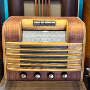 Op de afbeelding: Een RCA Victor radio met een houten behuizing in lichte en donkere houttinten. De voorzijde bevat een horizontale frequentieweergave, vijf zwarte drukknoppen, een luidsprekerrooster met vier horizontale lamellen en vier ronde draaiknoppen.