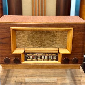 May include: A rectangular wooden radio featuring a dark walnut-toned veneer casing, a central beige fabric speaker grille, and a horizontal glass tuning dial with black numbers ranging from 55 to 160. Four circular brown control knobs are positioned below.