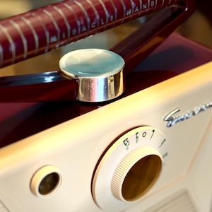 May include: Vintage Emerson radio with a cream-colored body and a burgundy handle. The handle has the text "MIRACLE WAND" in white. The radio features a tuning dial and a silver-colored button.