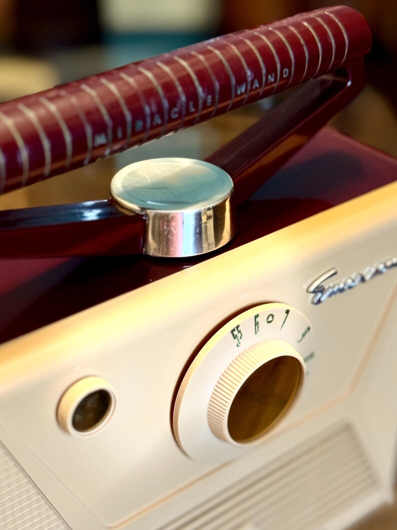 May include: Vintage Emerson radio with a cream-colored body and a burgundy handle. The handle has the text "MIRACLE WAND" in white. The radio features a tuning dial and a silver-colored button.