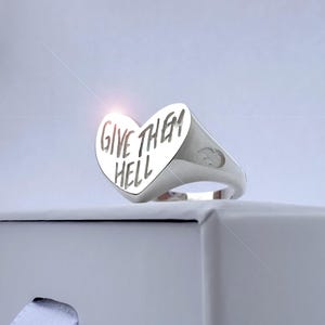 Peut inclure: Bague en argent avec une face en forme de cœur. La bague porte l'inscription "GIVE THEM HELL" en caractères gras sans empattement. La bague est posée sur un fond blanc.