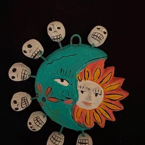 May include: A Day of the Dead-themed ornament featuring a turquoise crescent moon and a smiling sun. The sun is orange and yellow, and the moon has a face and is surrounded by eight white skull charms. The ornament has a loop for hanging.
