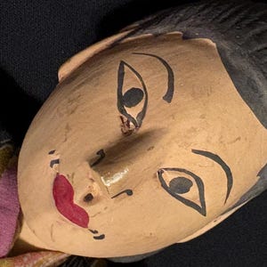 May include: A close-up of a painted doll's face with black hair and painted facial features. The doll's skin is tan, with black eyebrows, eyelashes, and a red mouth. The doll is wearing a colorful patterned garment.