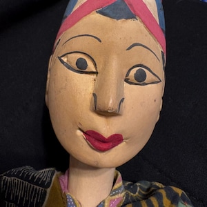 May include: A hand-carved wooden puppet head with painted features. The puppet has a light skin tone, black outlined eyes, and red lips. It wears a pink and blue headdress and a patterned fabric shirt.