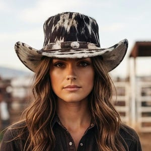 Può includere: Un cappello da cowboy con un motivo stampato a pelle di mucca bianco e nero. Il cappello presenta una corona strutturata e una tesa curva, accentuata da una fascia scura attorno alla base che include un piccolo ornamento metallico argentato sul davanti.