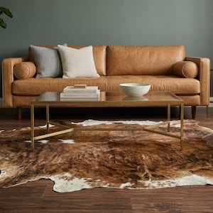 Può includere: Un tappeto in pelle bovina marrone e bianco dalla forma irregolare, caratterizzato da un motivo naturale maculato con chiazze marrone scuro, marrone chiaro e bianco, steso su un pavimento in legno scuro davanti a un divano in pelle beige.