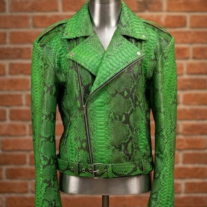 May include: A green snakeskin-patterned leather jacket with a notched collar, asymmetrical zipper closure, and a belt at the waist. The jacket is displayed on a mannequin against a brick wall background. The jacket has a classic biker style.