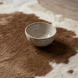 Puede incluir: Un cuenco peque&ntilde;o y redondo de cer&aacute;mica con un esmalte blanquecino moteado. El cuenco est&aacute; sobre una alfombra de piel de vaca marr&oacute;n y blanca. El cuenco tiene un dise&ntilde;o sencillo y minimalista.