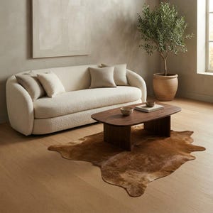 Puede incluir: Un sofá curvo de color crema con tapicería de tela texturizada, que incluye cuatro cojines cuadrados. Una mesa de centro de madera marrón oscuro con tablero rectangular redondeado se asienta sobre una alfombra de piel de vaca marrón, con dos cuencos y un libro.