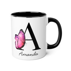 Peut inclure: Mug en céramique blanche avec une anse et un bord noirs. La tasse présente un grand « A » noir avec un motif de papillon rose et violet. Le nom « Amanda » est écrit en cursive en dessous.