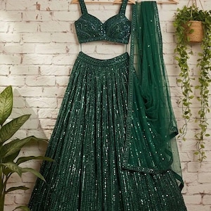 May include: Emerald green sequined lehenga set, including a crop top with wide straps, a full skirt, and a matching sheer dupatta. The outfit hangs on wooden hangers against a white brick wall.