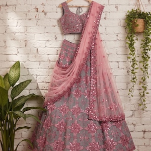 Könnte beinhalten: Ein graues Lehenga-Choli-Set bestehend aus einem ärmellosen Oberteil und einem ausgestellten Rock, beide verziert mit aufwendigen rosa Blumenstickereien und reflektierenden Pailletten. Ein passender, transparenter rosa Netz-Dupatta liegt über dem Rock.