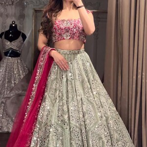 A person wearing a dark red embroidered choli, a light green lehenga skirt with silver embroidery and mirrors, and a hot pink net dupatta, standing in front of a mirror.