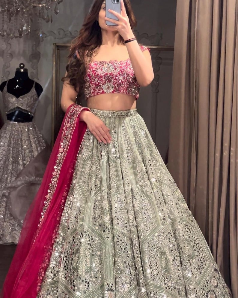 A person wearing a dark red embroidered choli, a light green lehenga skirt with silver embroidery and mirrors, and a hot pink net dupatta, standing in front of a mirror.