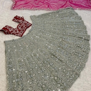 A flat lay of a light green lehenga skirt with silver embroidery and mirrors, a dark red embroidered choli, and a hot pink net dupatta, on a white textured surface.