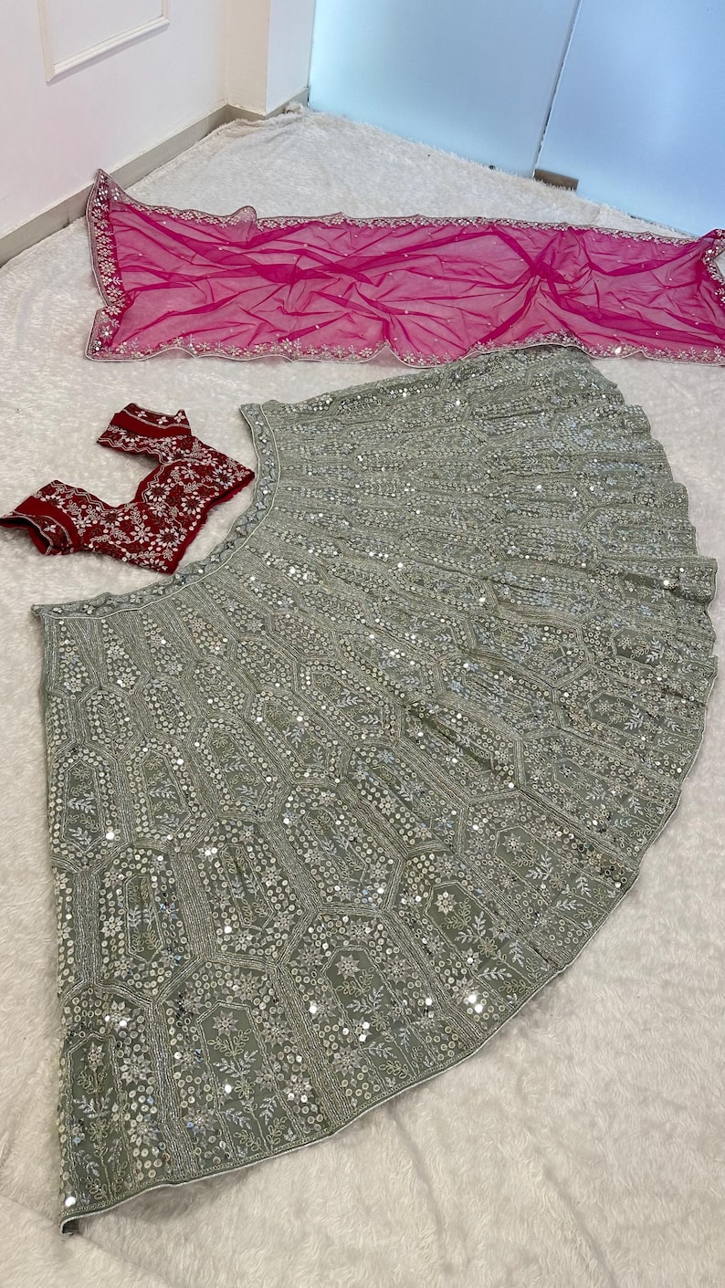 A flat lay of a light green lehenga skirt with silver embroidery and mirrors, a dark red embroidered choli, and a hot pink net dupatta, on a white textured surface.