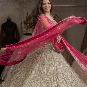 A person with long brown hair, wearing a light green lehenga skirt with silver embroidery and mirrors, a dark red embroidered choli, and a hot pink net dupatta, spinning in a room with a chandelier.