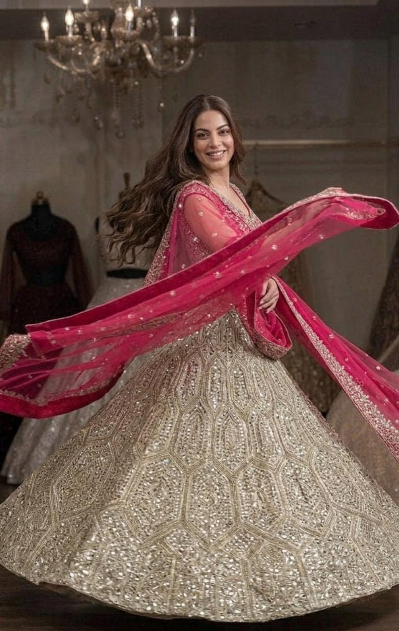 A person with long brown hair, wearing a light green lehenga skirt with silver embroidery and mirrors, a dark red embroidered choli, and a hot pink net dupatta, spinning in a room with a chandelier.