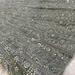 A close-up of a sage green fabric with intricate silver embroidery and sequins in floral and geometric patterns.