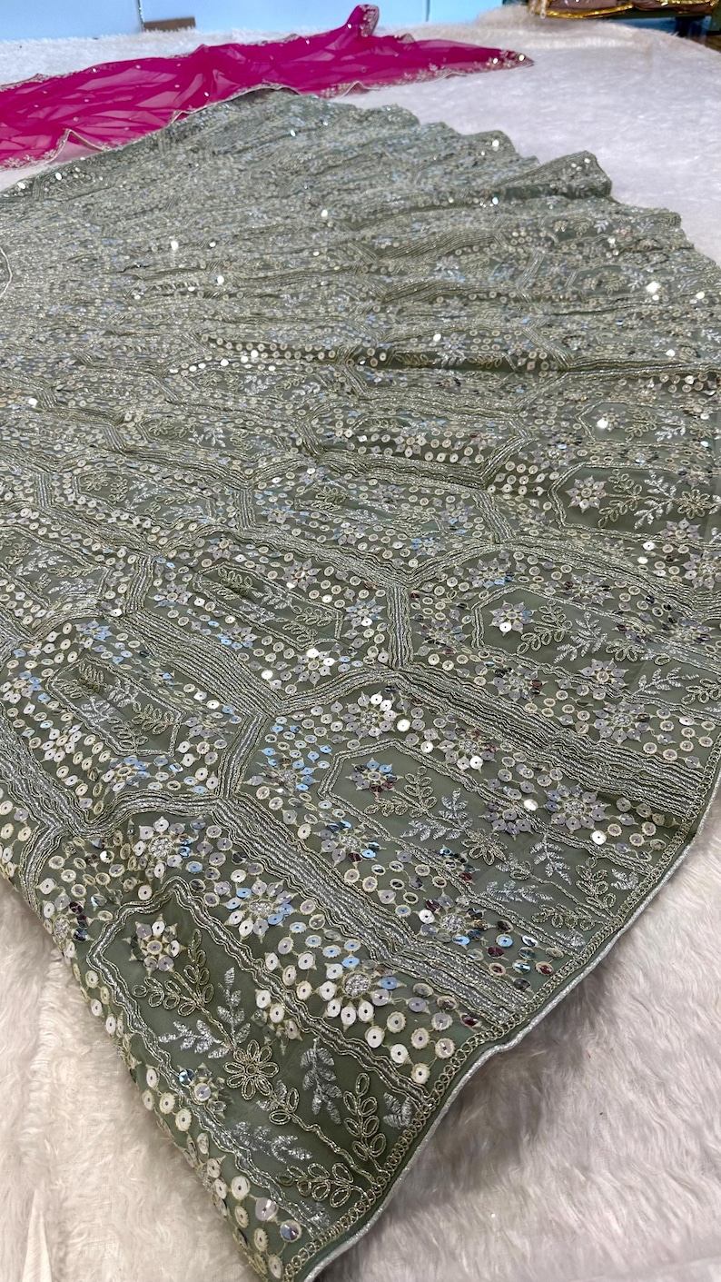 A close-up of a sage green fabric with intricate silver embroidery and sequins in floral and geometric patterns.