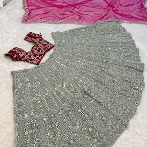 A flat lay of a light green lehenga skirt with silver embroidery and mirrors, a dark red embroidered choli, and a hot pink net dupatta, on a white textured surface.