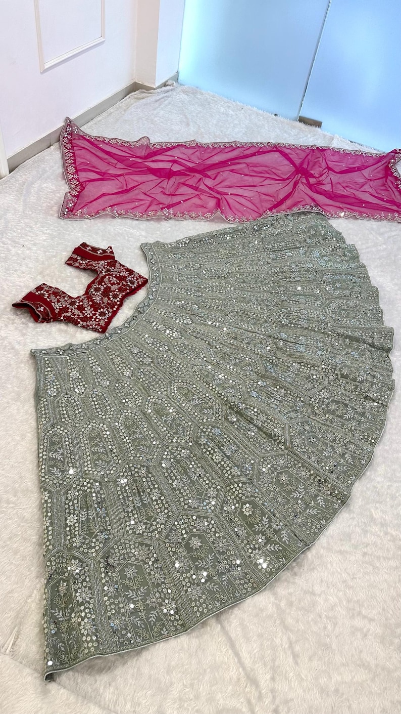 A flat lay of a light green lehenga skirt with silver embroidery and mirrors, a dark red embroidered choli, and a hot pink net dupatta, on a white textured surface.