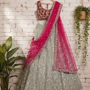 A light green lehenga skirt with silver embroidery and mirrors, a dark red embroidered choli, and a hot pink net dupatta with silver trim, hanging on a hanger against a white brick wall.