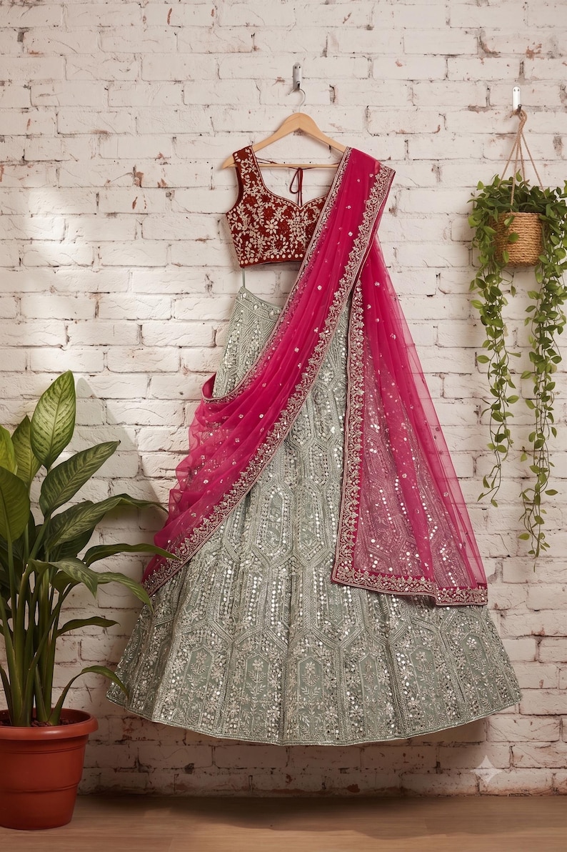 A light green lehenga skirt with silver embroidery and mirrors, a dark red embroidered choli, and a hot pink net dupatta with silver trim, hanging on a hanger against a white brick wall.