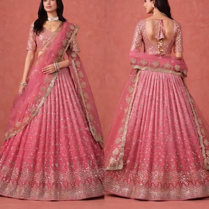 Peut inclure: Un ensemble lehenga choli rose composé d'un chemisier à manches courtes et d'une jupe plissée volumineuse, ornés de broderies argentées et de sequins. Un dupatta transparent assorti avec une bordure décorative complète la tenue.