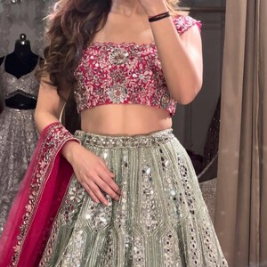 A person from the chest down, wearing a dark red off-the-shoulder choli with silver embroidery and a light green lehenga skirt with silver embroidery and mirrors, with a hot pink dupatta draped over their arm.