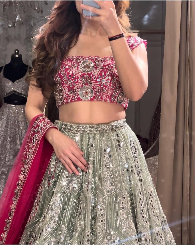 A person from the chest down, wearing a dark red off-the-shoulder choli with silver embroidery and a light green lehenga skirt with silver embroidery and mirrors, with a hot pink dupatta draped over their arm.
